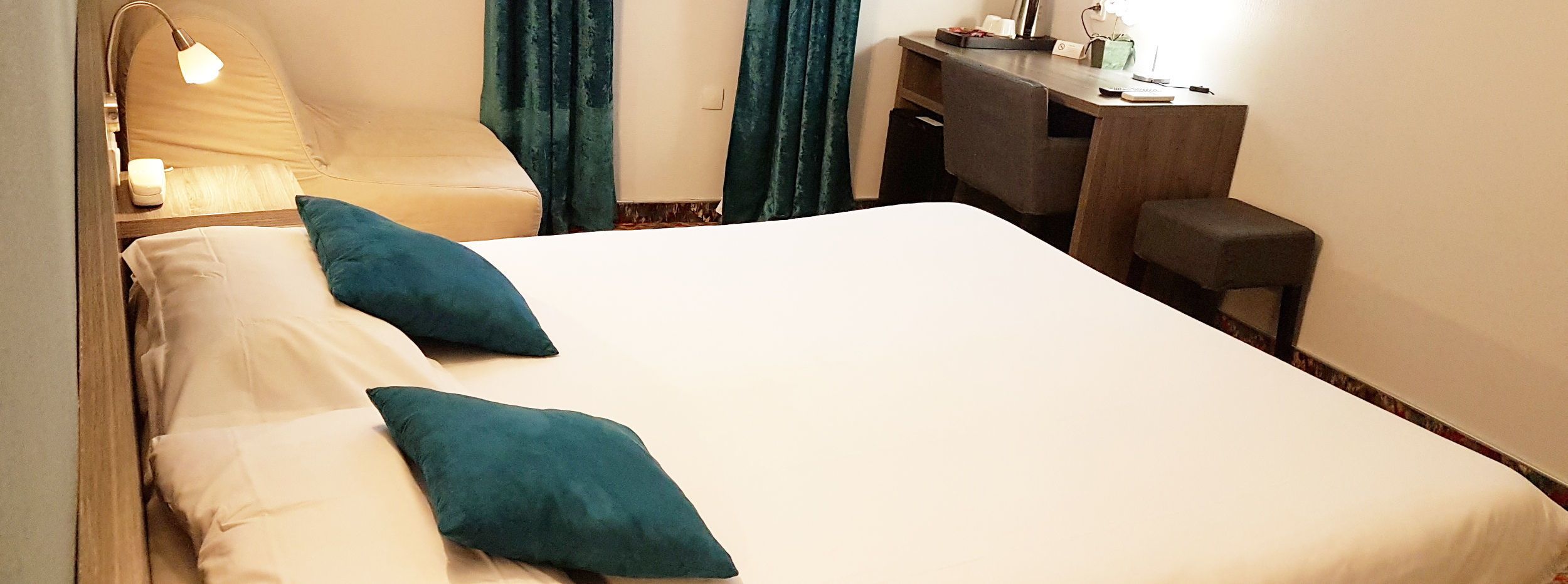 Une chambre d’hôtel avec un lit, deux oreillers, un bureau, une chaise, une lampe et une fenêtre avec des rideaux.