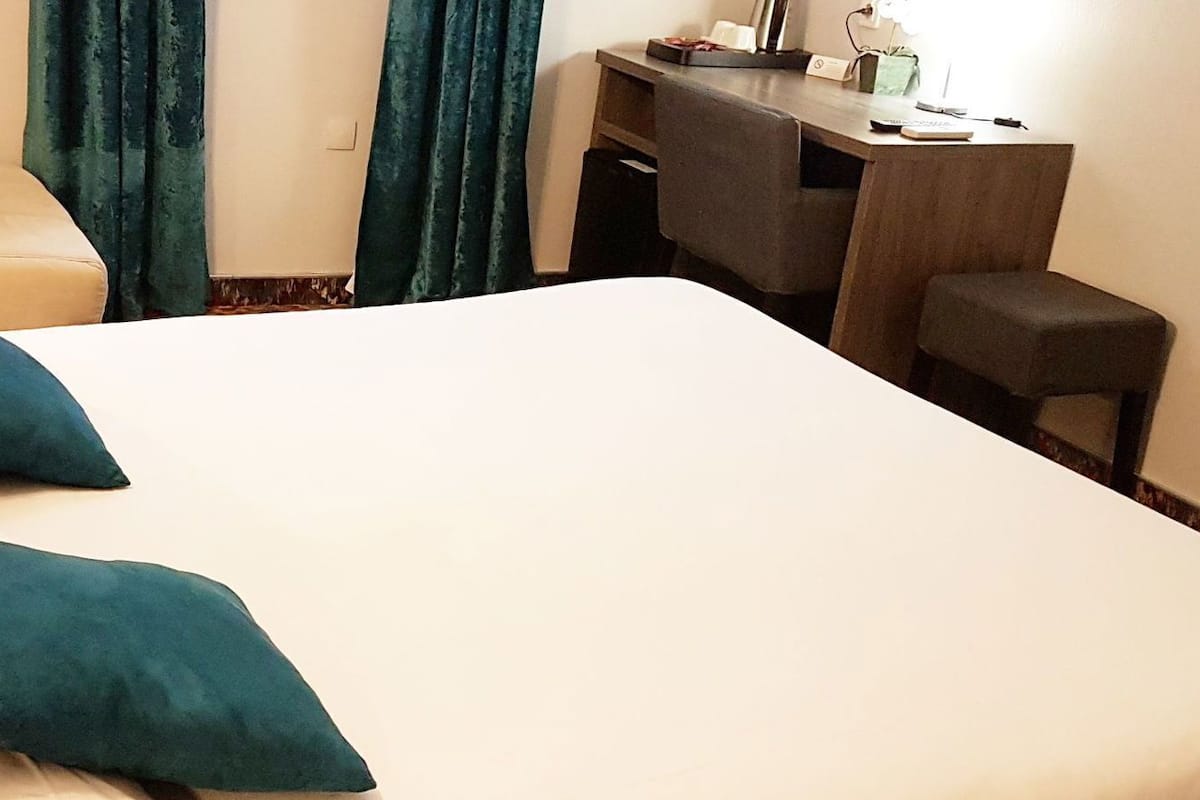 A hotel room with a bed, two pillows, a desk, a chair, a lamp, and curtains.