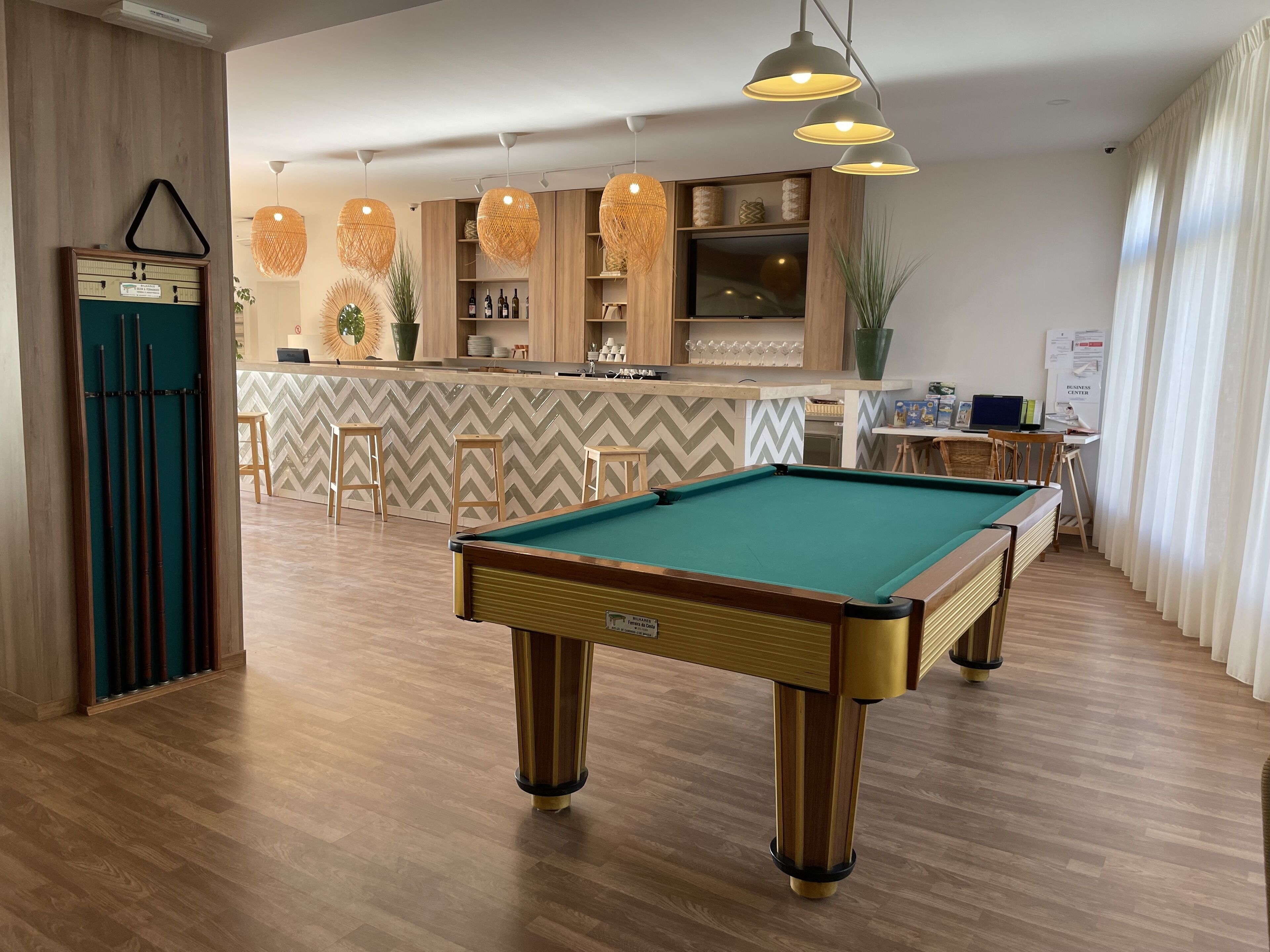 A pool table in a well-lit room with wooden flooring, a bar area with shelves, and a business center.