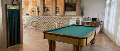 A pool table in a well-lit room with wooden flooring, a bar area with shelves, and a business center.