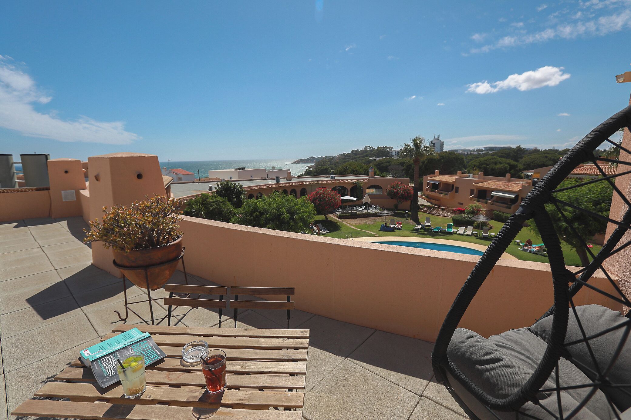 A terrace with a wooden table, a chair, a potted plant, and a view of a swimming pool and buildings in the distance.