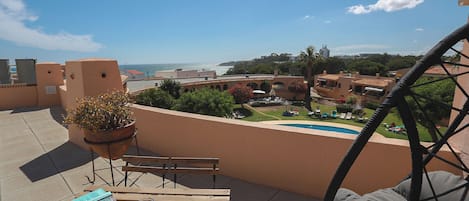 A terrace with a wooden table, a chair, a potted plant, and a view of a swimming pool and buildings in the distance.