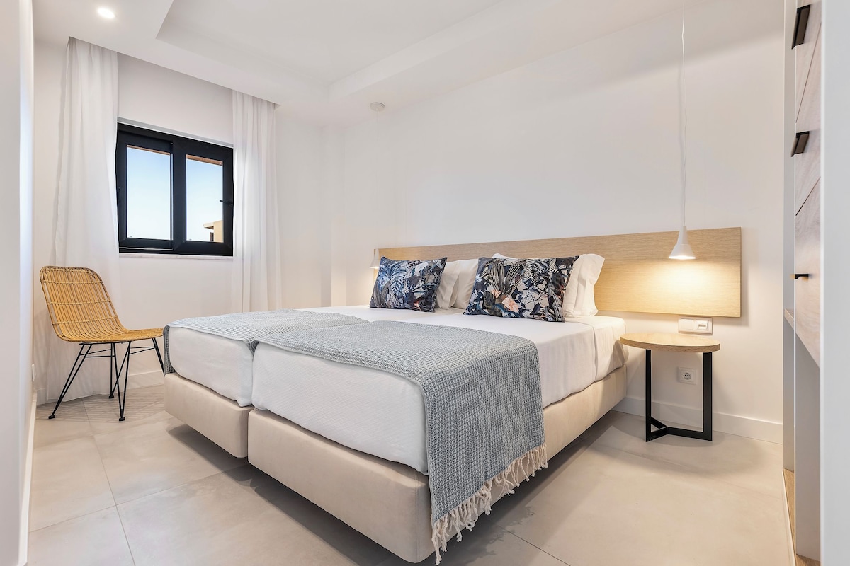 Un dormitorio moderno con una cama grande, una mesita de noche con lámpara, una silla y una ventana con cortinas.