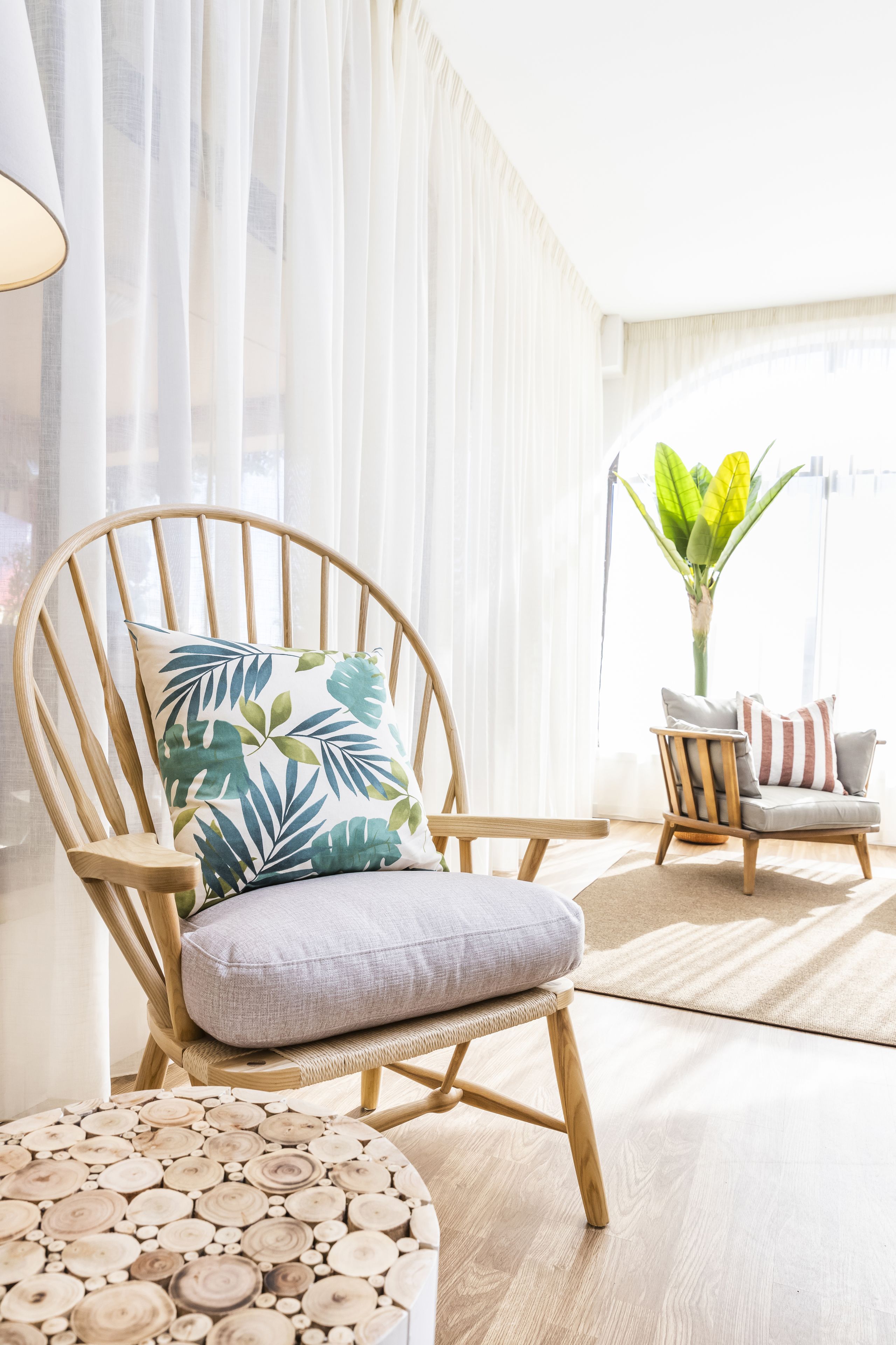 Una habitación con una silla de madera, un cojín estampado con motivos de hojas, una mesa redonda de madera y una planta en maceta.