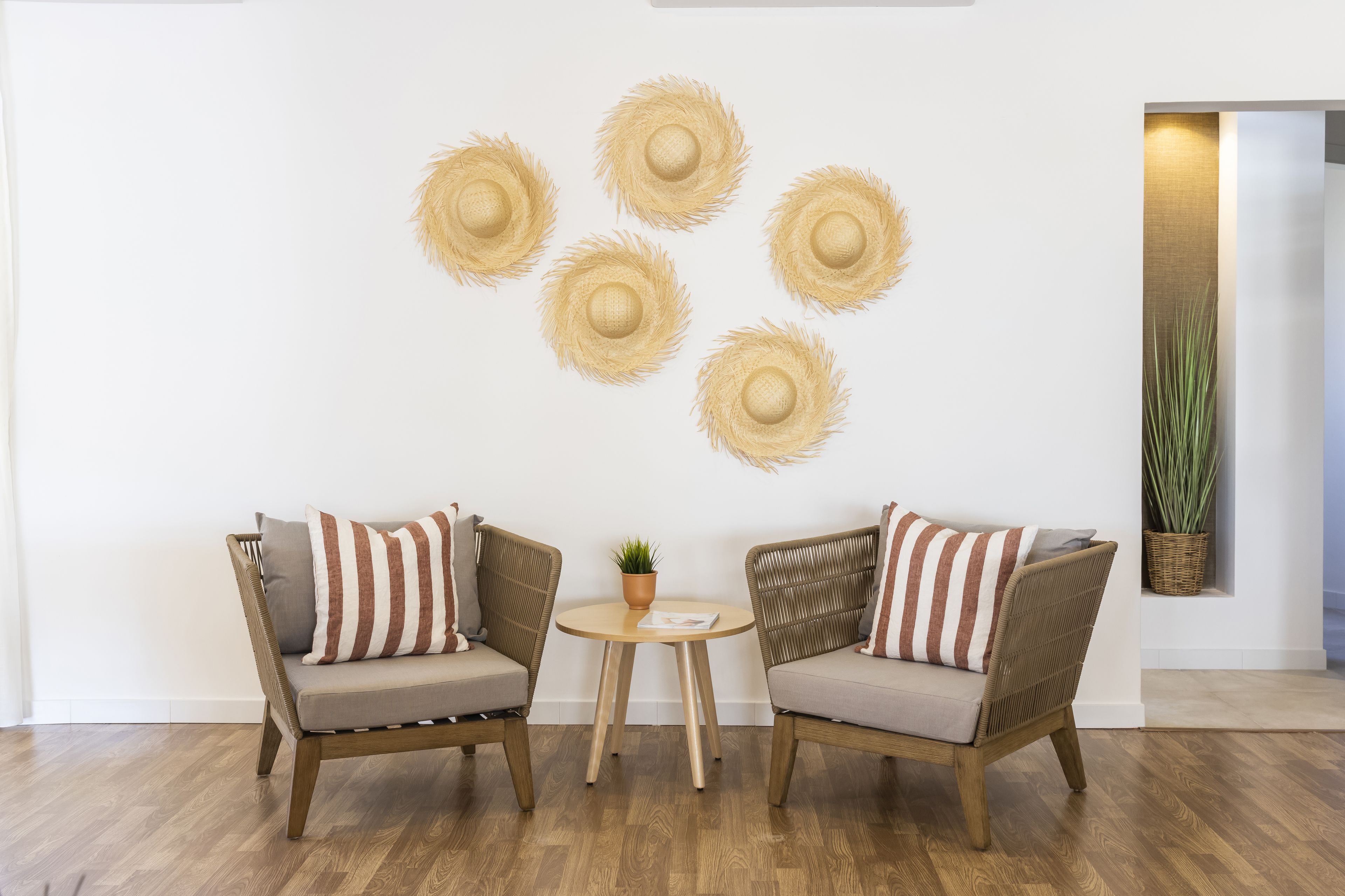 Una habitación con dos sillas de mimbre, una mesita redonda con una planta y un muro decorado con cinco sombreros de paja.