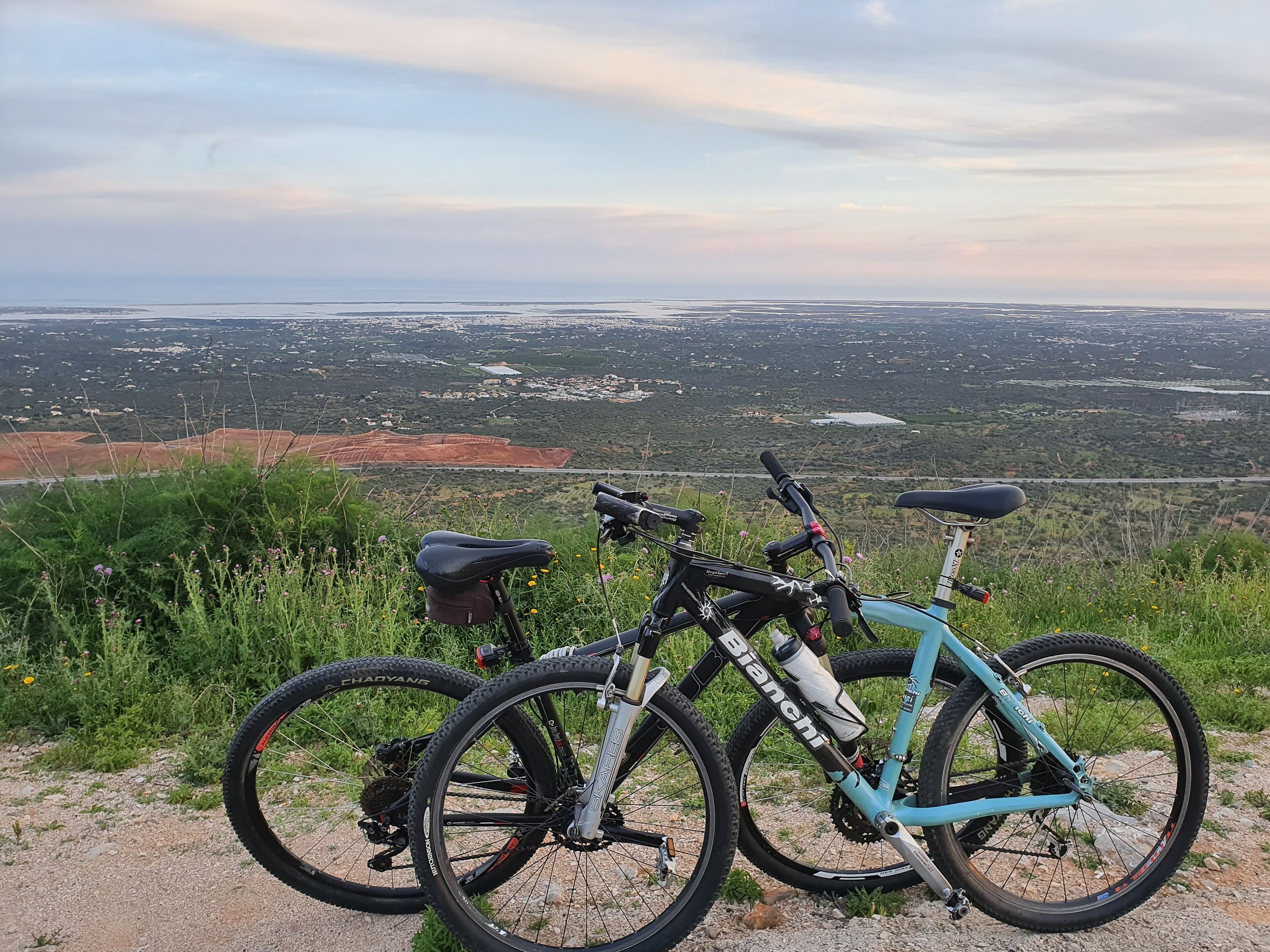 Due biciclette parcheggiate su una collina erbosa con vista su una valle e una città in lontananza, oltre a un'ampia zona aperta.