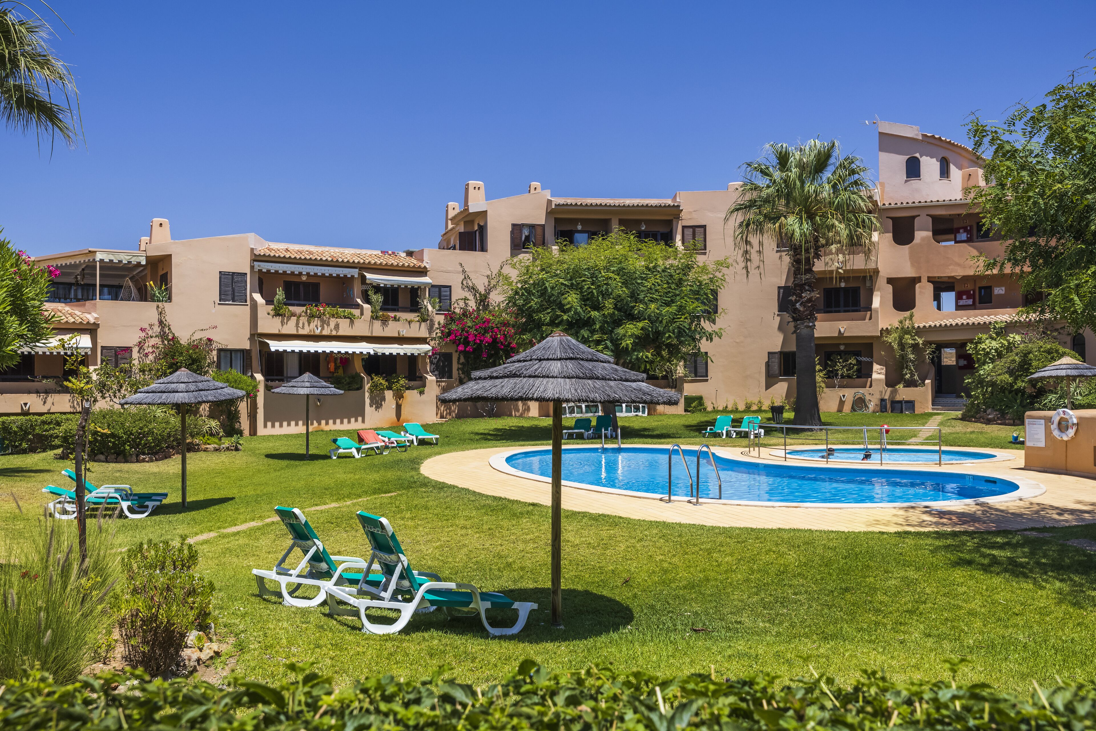 A pool area with lounge chairs and umbrellas, surrounded by buildings with balconies and a clear blue sky.