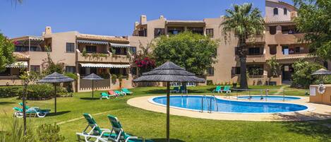 A pool area with lounge chairs and umbrellas, surrounded by buildings with balconies and a clear blue sky.