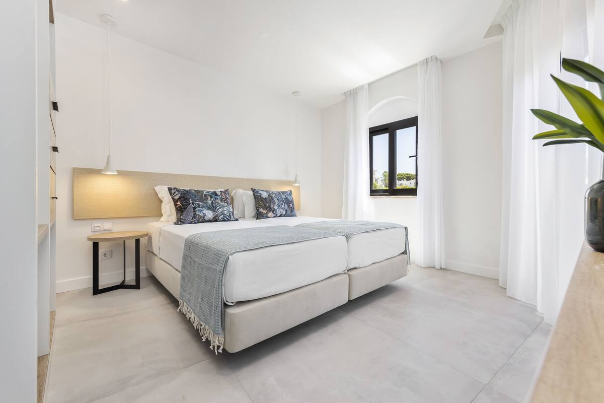 Quarto moderno com uma cama grande, mesas de cabeceira, um criado-mudo, uma janela com cortinas e uma planta em vaso.