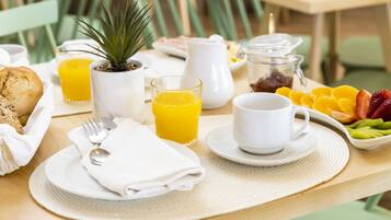 Un desayuno con tostadas, un vaso de zumo de naranja, una taza blanca, un bol de fruta y una pequeña planta en maceta sobre una mesa.