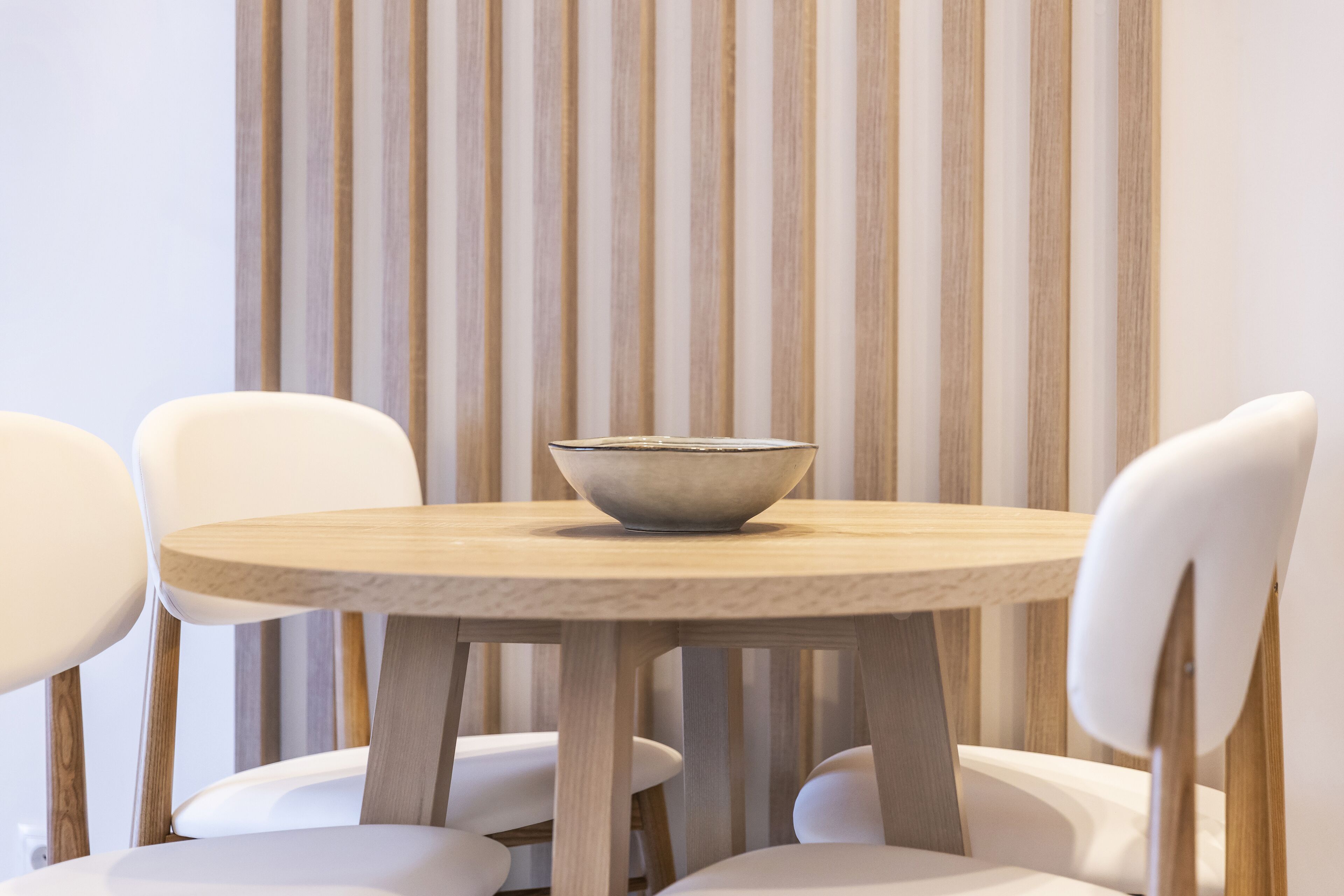 Una mesa redonda de madera con un cuenco en el centro, rodeada por cuatro sillas blancas.