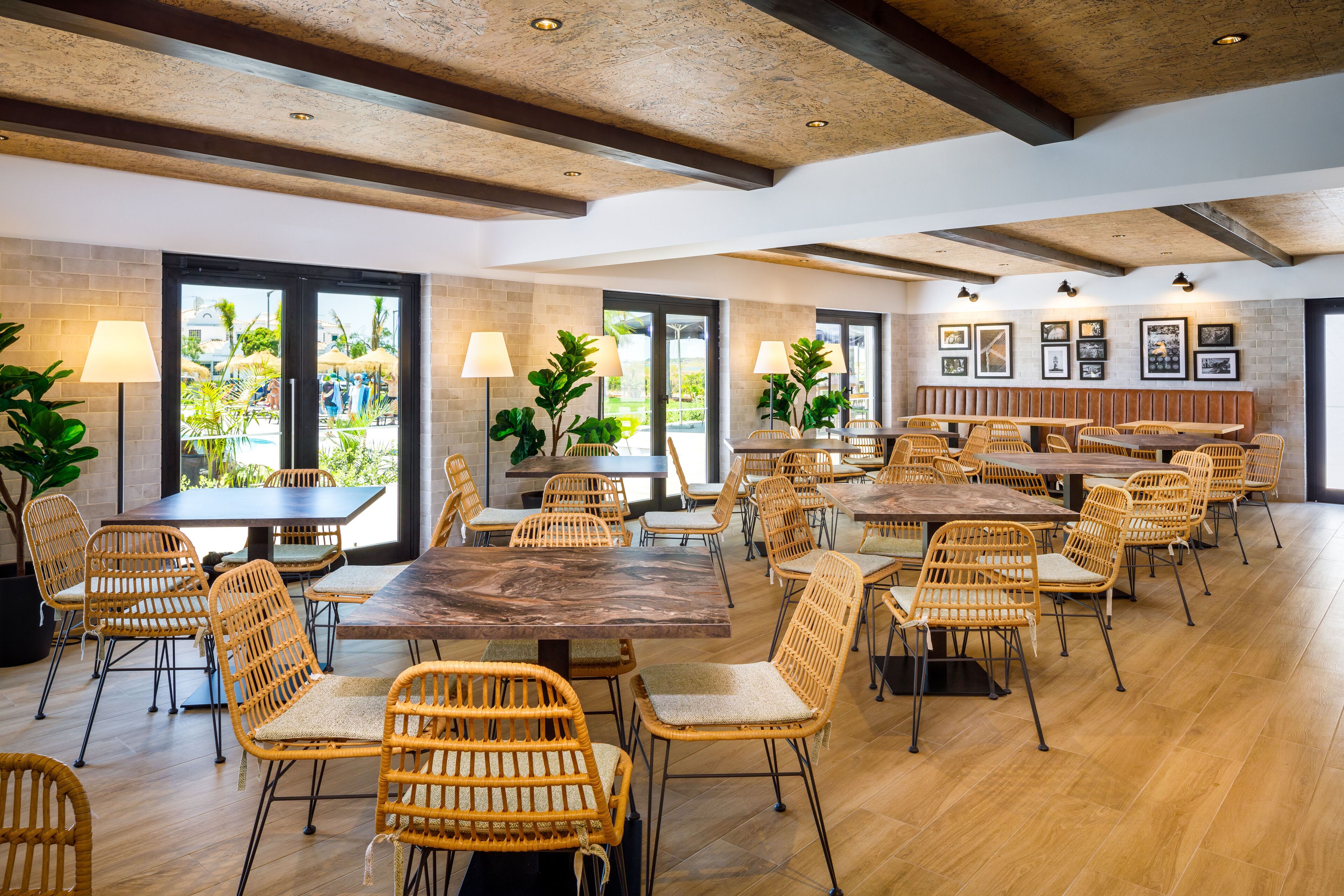 A spacious dining area with multiple tables and wicker chairs, large windows with views of greenery, and a rustic ceiling design.