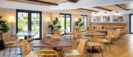 A spacious dining area with multiple tables and wicker chairs, large windows with views of greenery, and a rustic ceiling design.