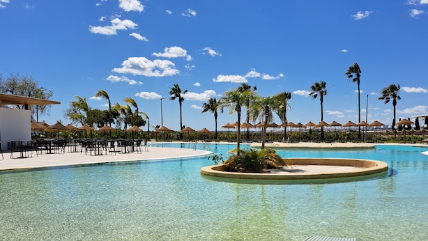 A clear pool with lounge chairs, palm trees, and a white building in the background. - AP Cabanas Beach & Nature - Adults Friendly (Tavira)