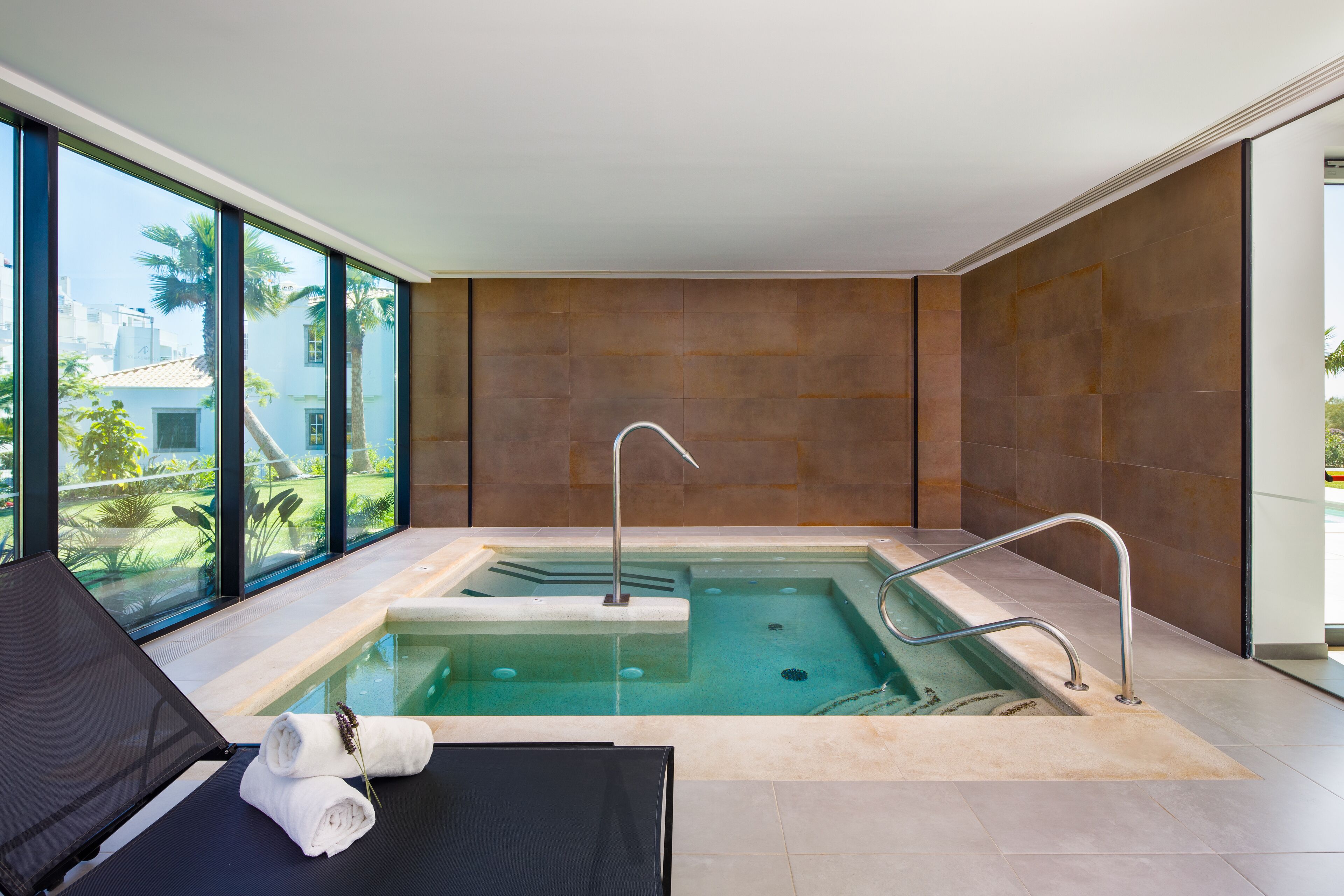 Un espace piscine intérieur moderne, doté d’un fauteuil de relaxation, d’un petit jacuzzi et de grandes fenêtres offrant une vue sur les palmiers et les bâtiments.