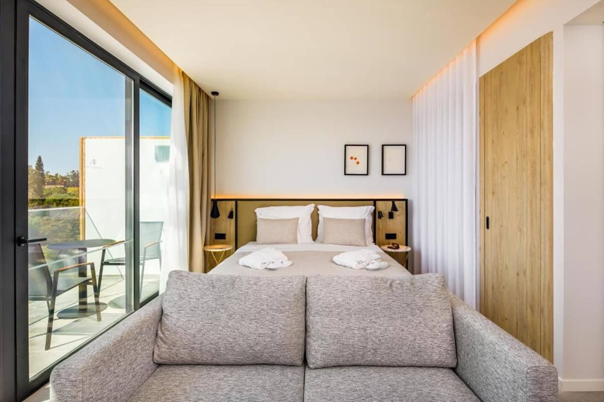 Et moderne hotelværelse med en stor seng, en sofa, en balkon med et bord og stole samt udsigt over byen.