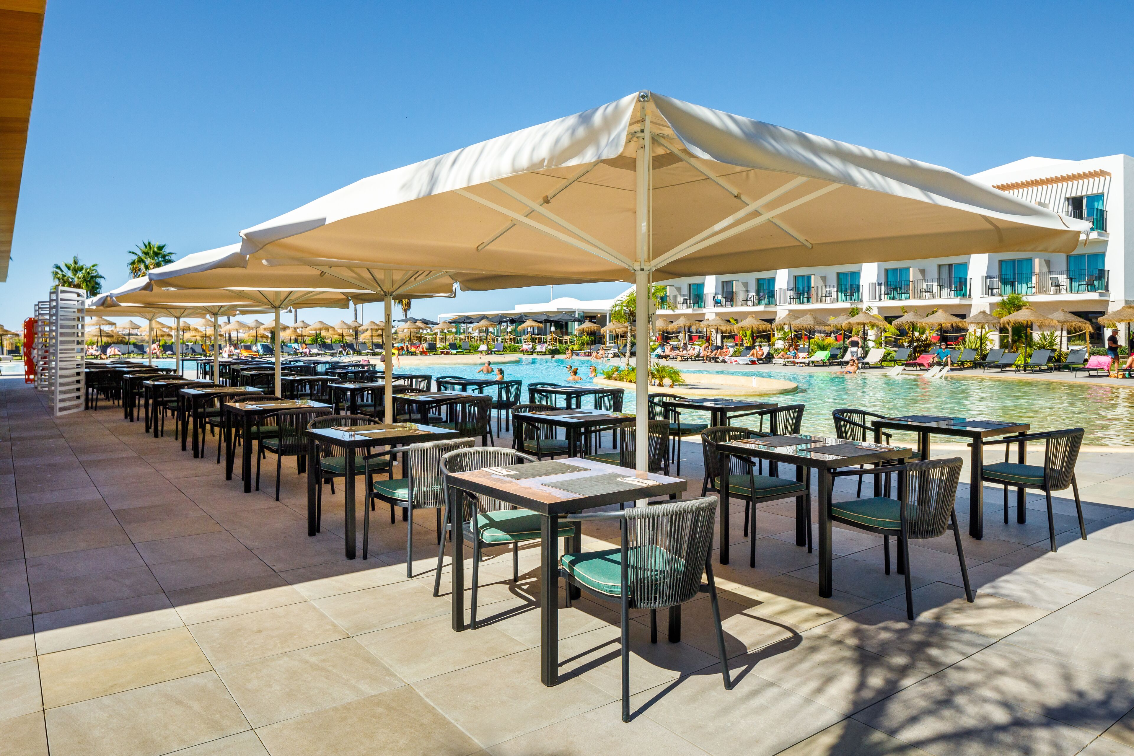 An outdoor pool area with multiple tables and chairs under large umbrellas, surrounded by a pool and a building with balconies.
