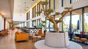 A spacious indoor area with a long row of tables and chairs, a large tree, and a modern architectural design with large windows. - AP Cabanas Beach & Nature - Adults Friendly (Tavira)