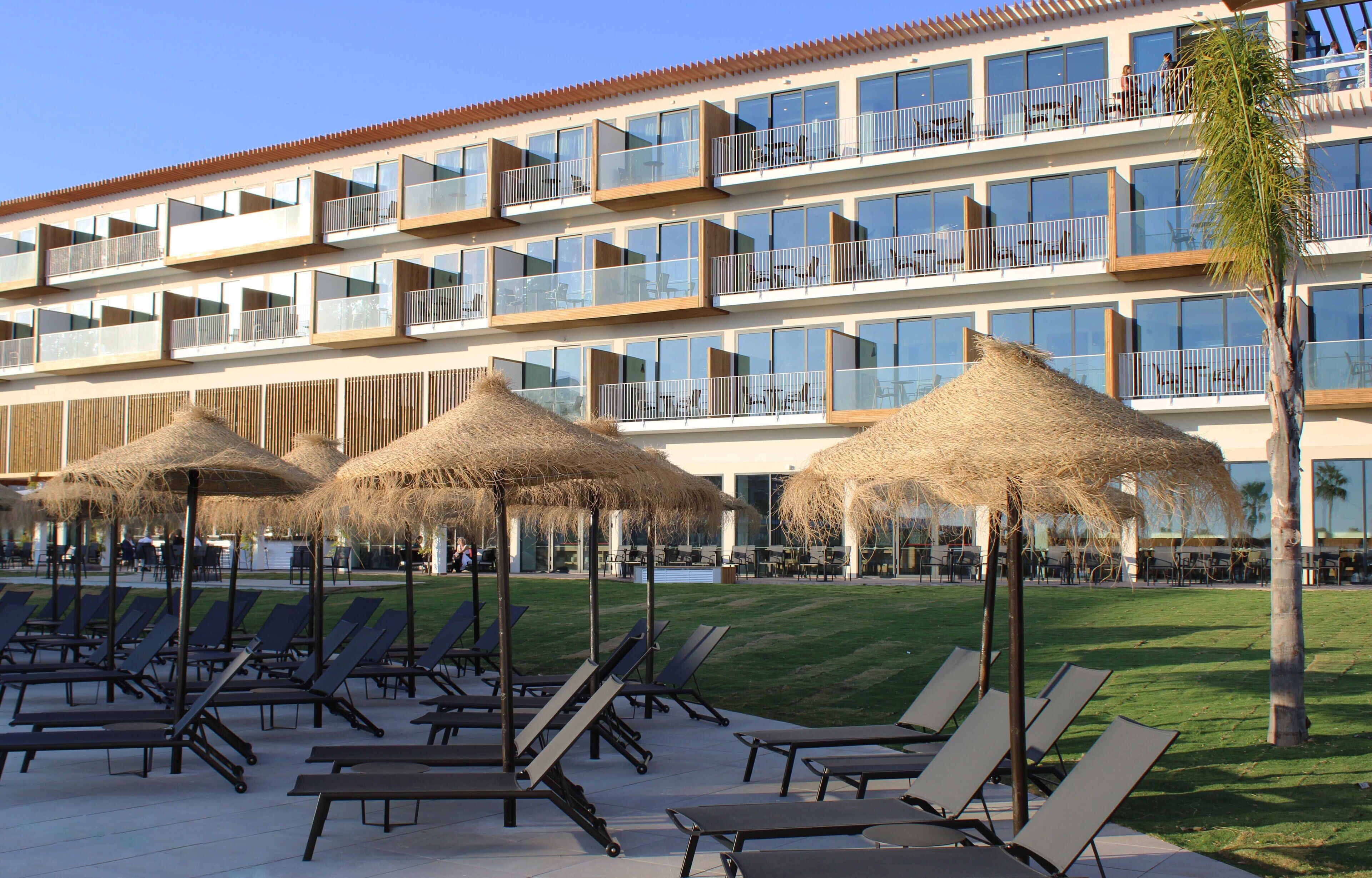 Un hotel con múltiples balcones, zonas de estar al aire libre con sombrillas de paja, y una piscina al fondo.