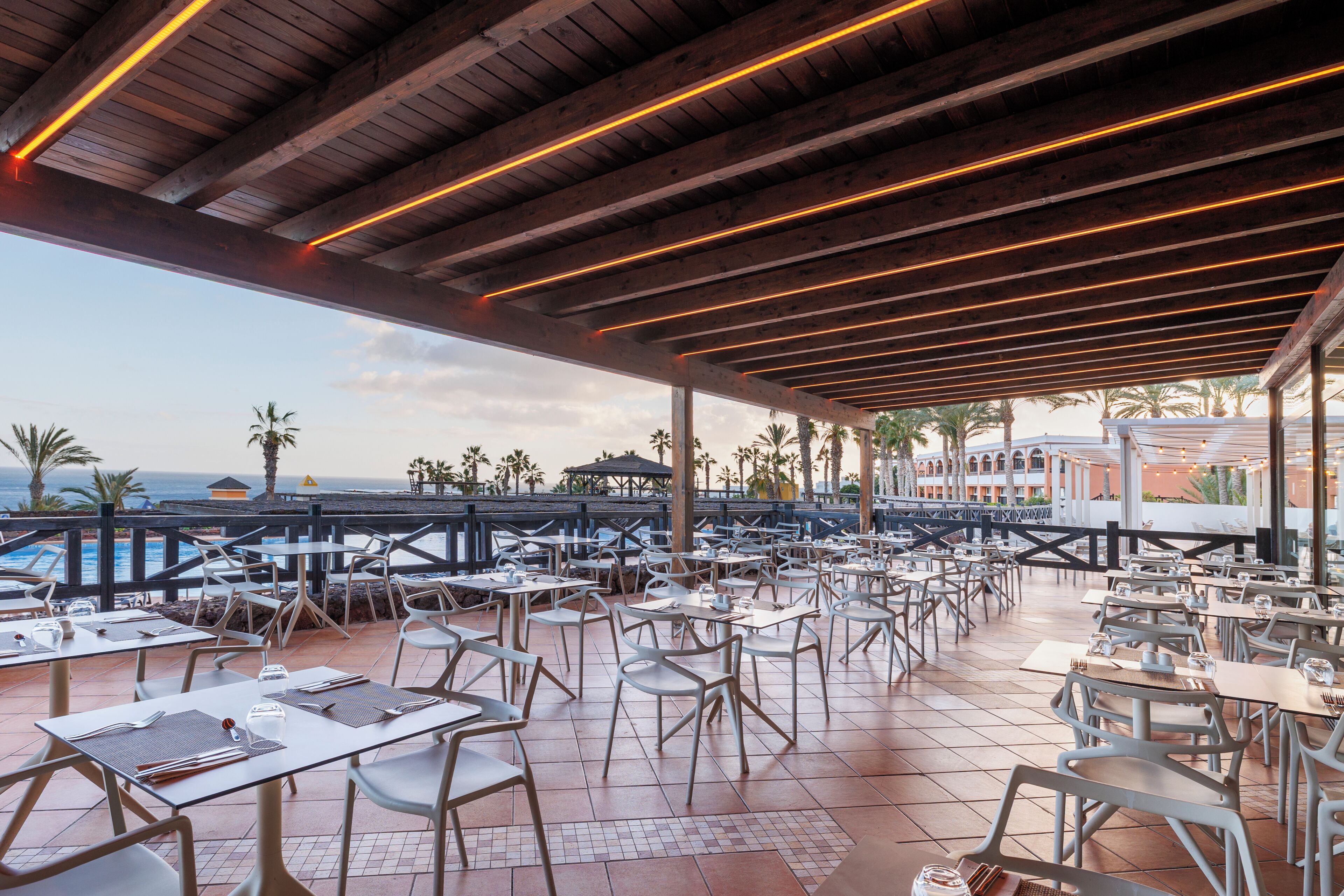 Un espace extérieur aménagé pour les repas, avec des tables et des chaises, offrant une vue sur une piscine et des palmiers.