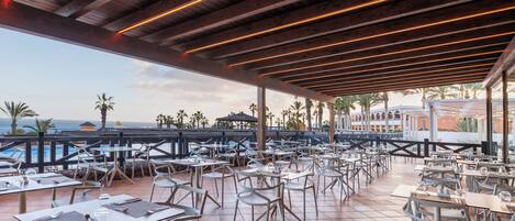 An outdoor dining area with tables and chairs, overlooking a pool and palm trees.