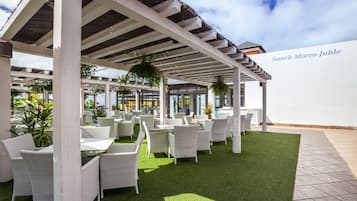 Área de estar externa com móveis de vime branco e pérgola branca.