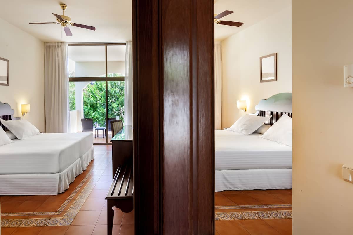 A hotel room with two beds, a ceiling fan, a view of greenery outside, and a dining area with a table and chairs.