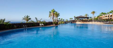 A large outdoor swimming pool with clear blue water, surrounded by palm trees and a gazebo.