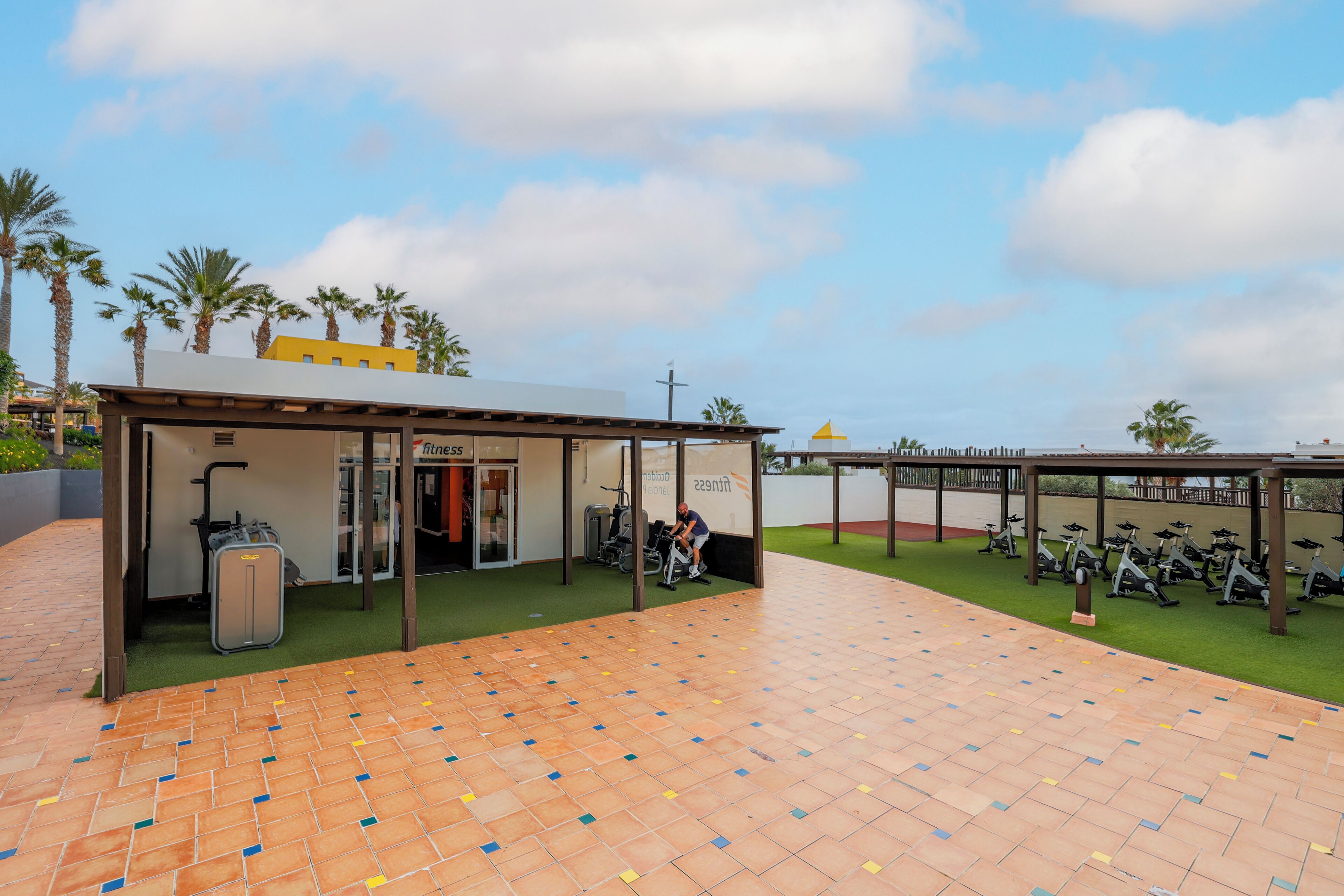 Un área de gimnasio al aire libre con bicicletas de ejercicio, una zona de estar cubierta y un carrito para equipaje.