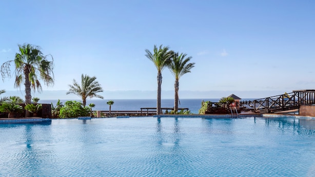A pool with a view of the ocean, surrounded by palm trees and a wooden deck. - Occidental Jandía Playa (Pajara)