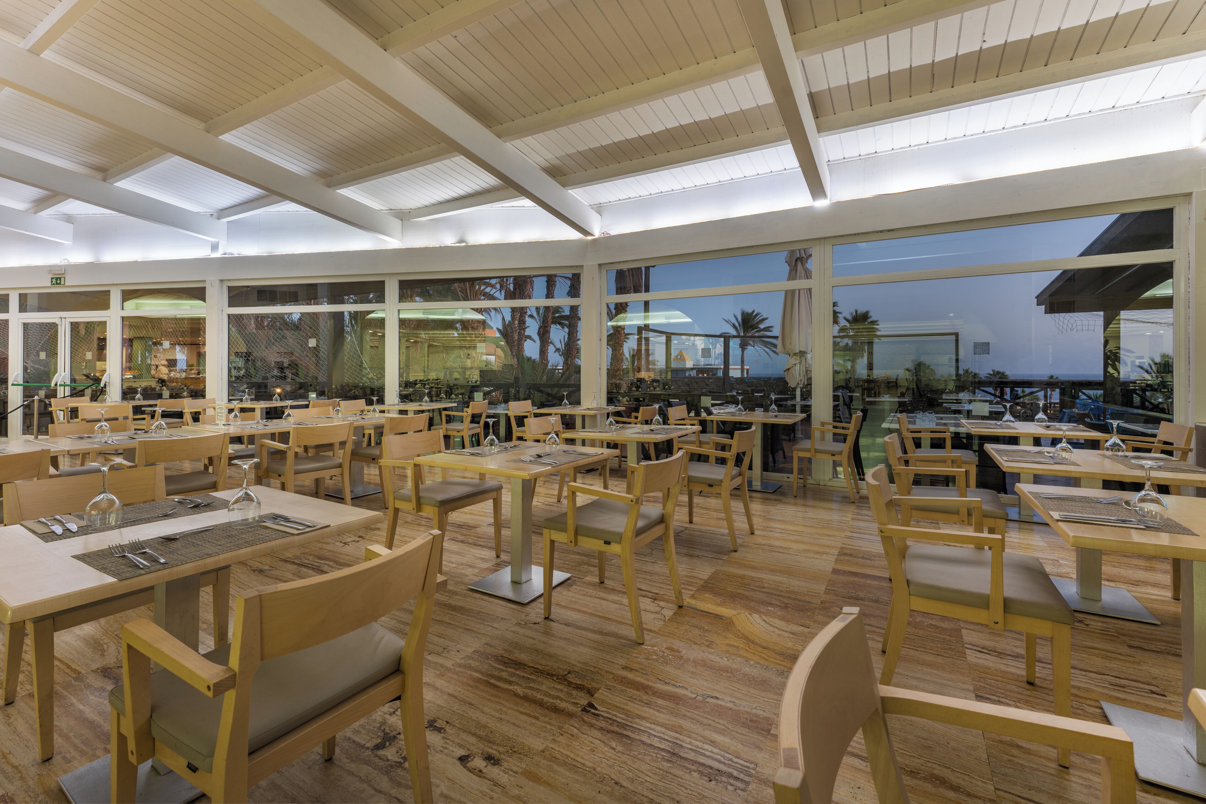 Un restaurante con mesas y sillas de madera, grandes ventanales y vistas a palmeras.
