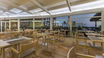 Un restaurante con mesas y sillas de madera, grandes ventanales y vistas a palmeras.
