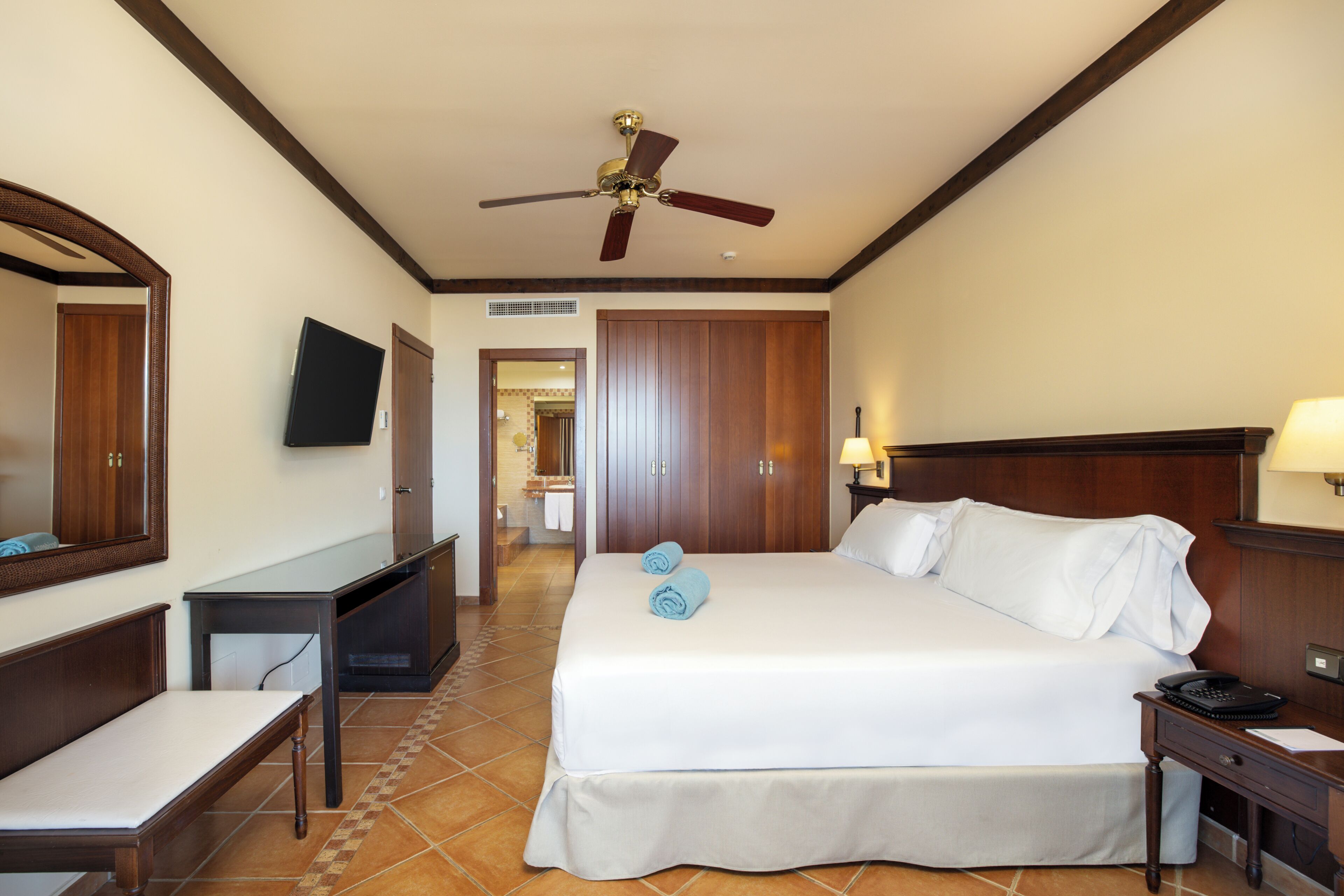 Une chambre d’hôtel avec un lit, un bureau, une chaise, une télévision, un ventilateur de plafond et une salle de bain visible au fond.