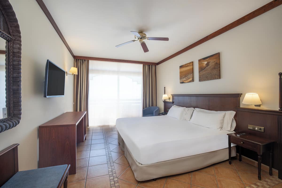 A hotel room with a large bed, a ceiling fan, a flat-screen TV, a desk, and a chair.