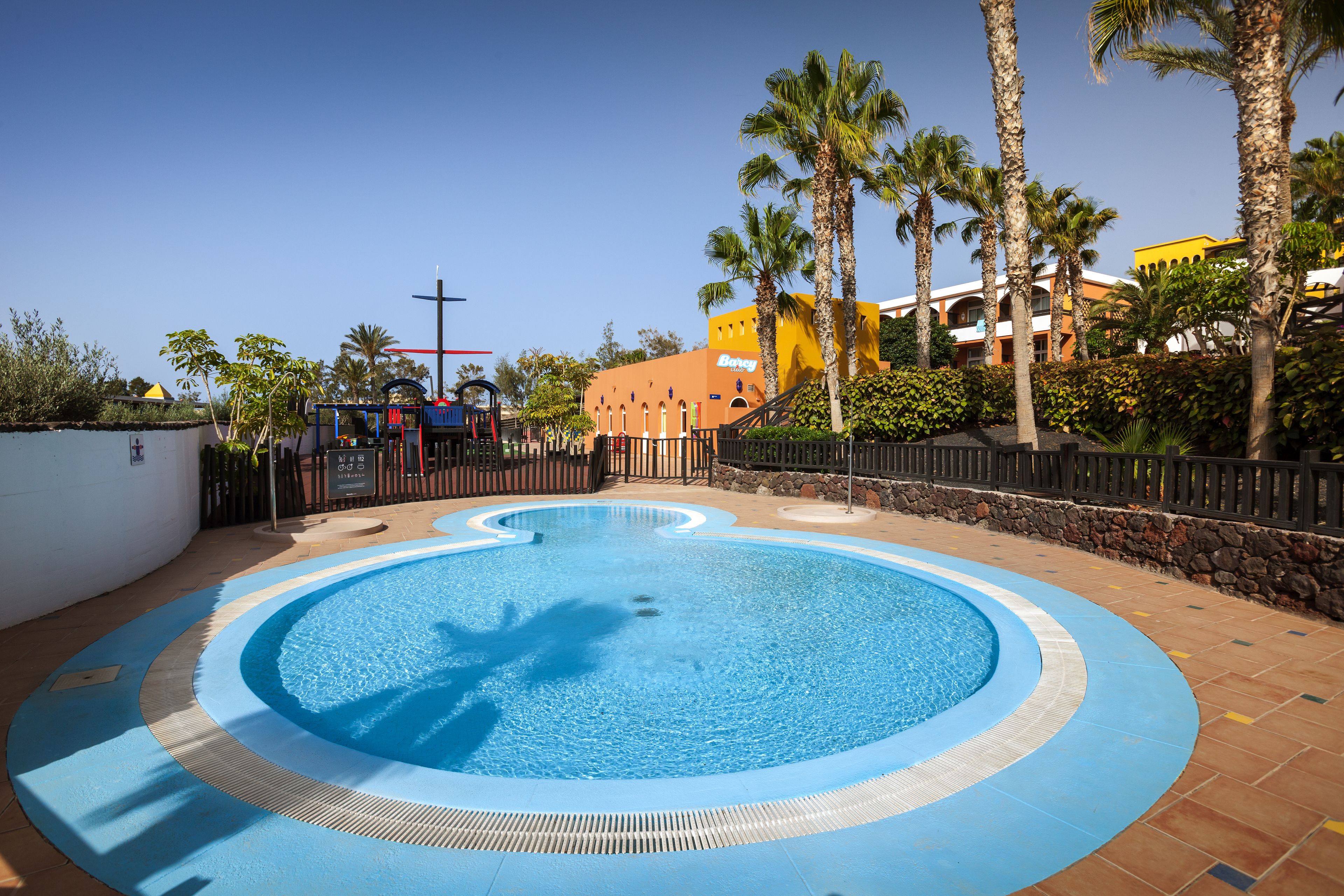 A swimming pool with a circular design, surrounded by palm trees and a building with a yellow facade.