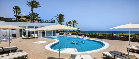 A pool area with lounge chairs and umbrellas, a clear view of the ocean, and a building with a balcony.