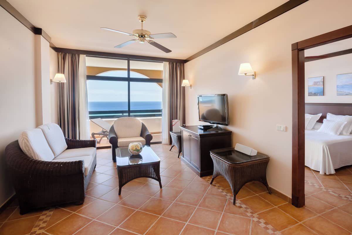 A hotel room with a bed, a television, a seating area with a chair and a table, a small desk, and a view of the sea through large windows.