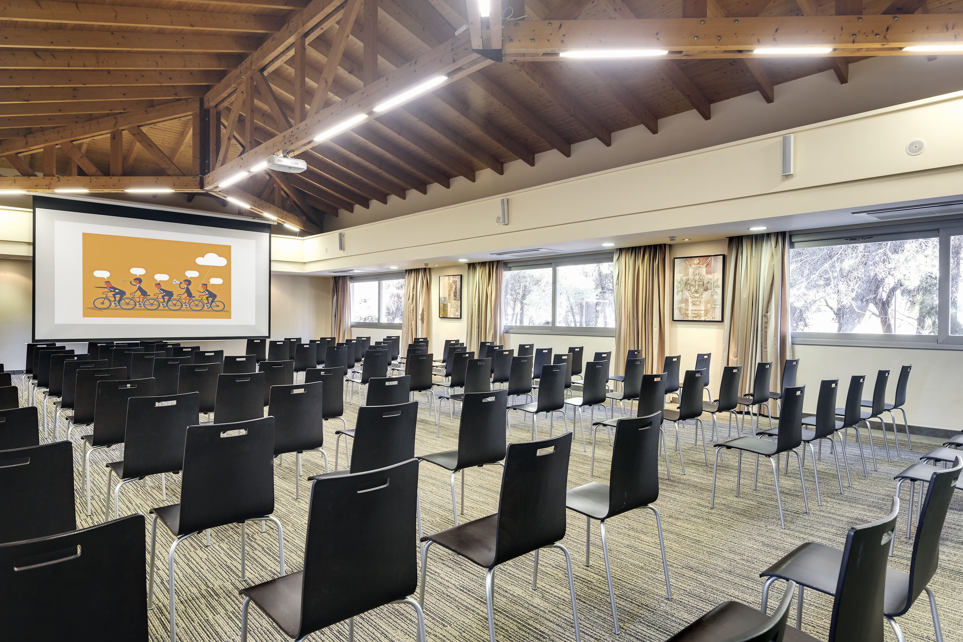 A conference room with rows of black chairs, a projector screen, and large windows with curtains.