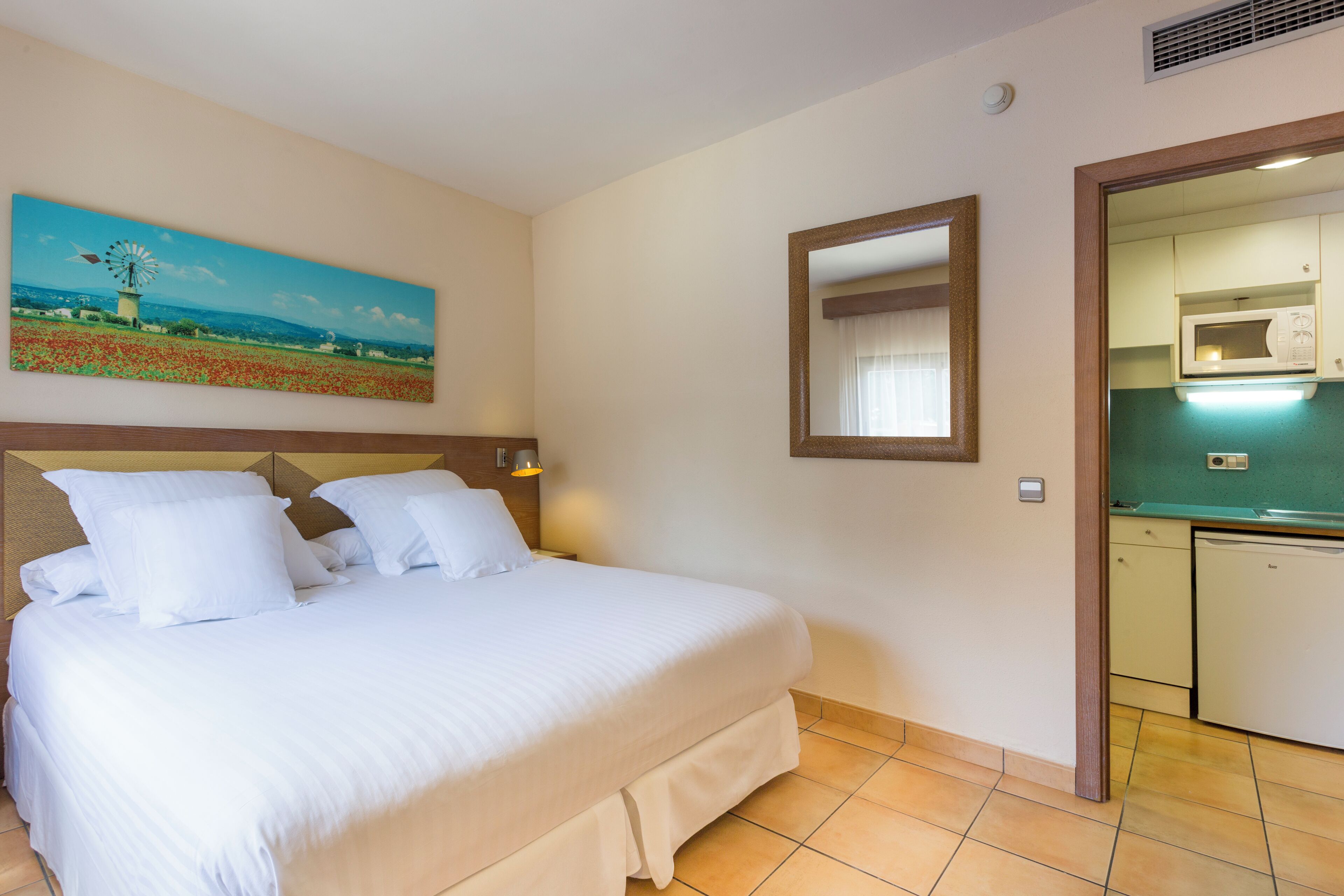 A hotel room with a large bed, a painting of a windmill, a mirror, and a kitchenette.
