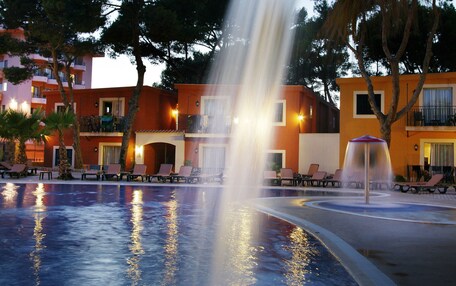 A swimming pool with a water fountain, lounge chairs, and a building with balconies in the background.
