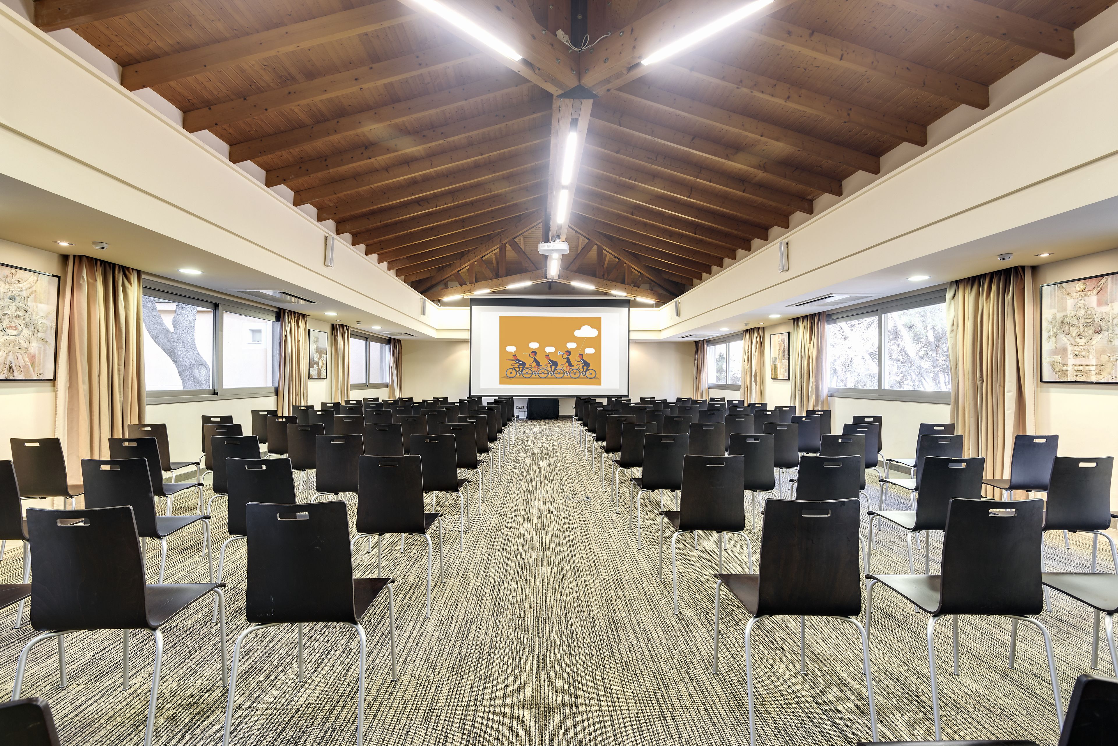 Un salón de conferencias con filas de sillas, vigas de madera en el techo y ventanas amplias.