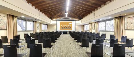 Un salón de conferencias con filas de sillas, vigas de madera en el techo y ventanas amplias.