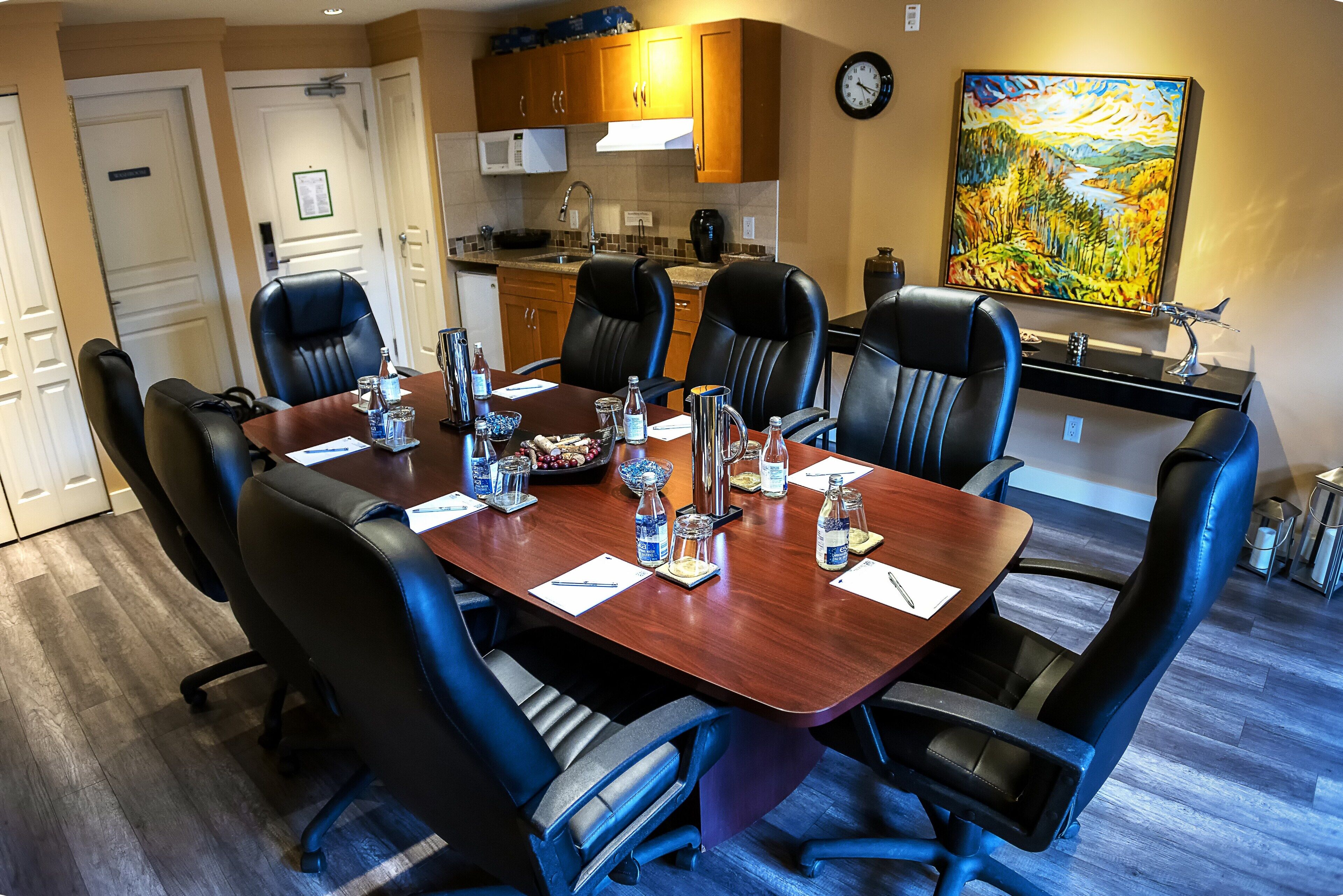 A conference room with a wooden table, black leather chairs, and a kitchen area in the background.