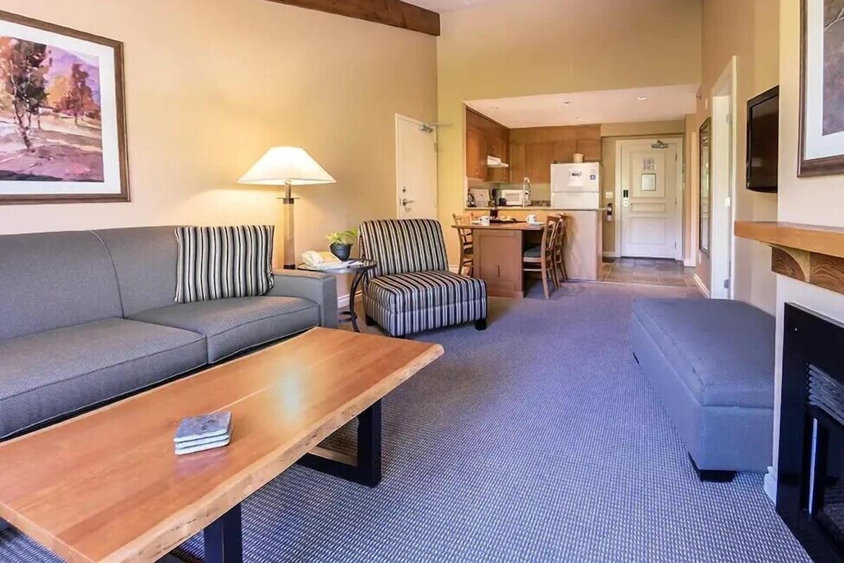 Una habitación de hotel moderna con sofá, sillón a rayas, mesa de centro de madera y una pequeña cocina con refrigerador y microondas.
