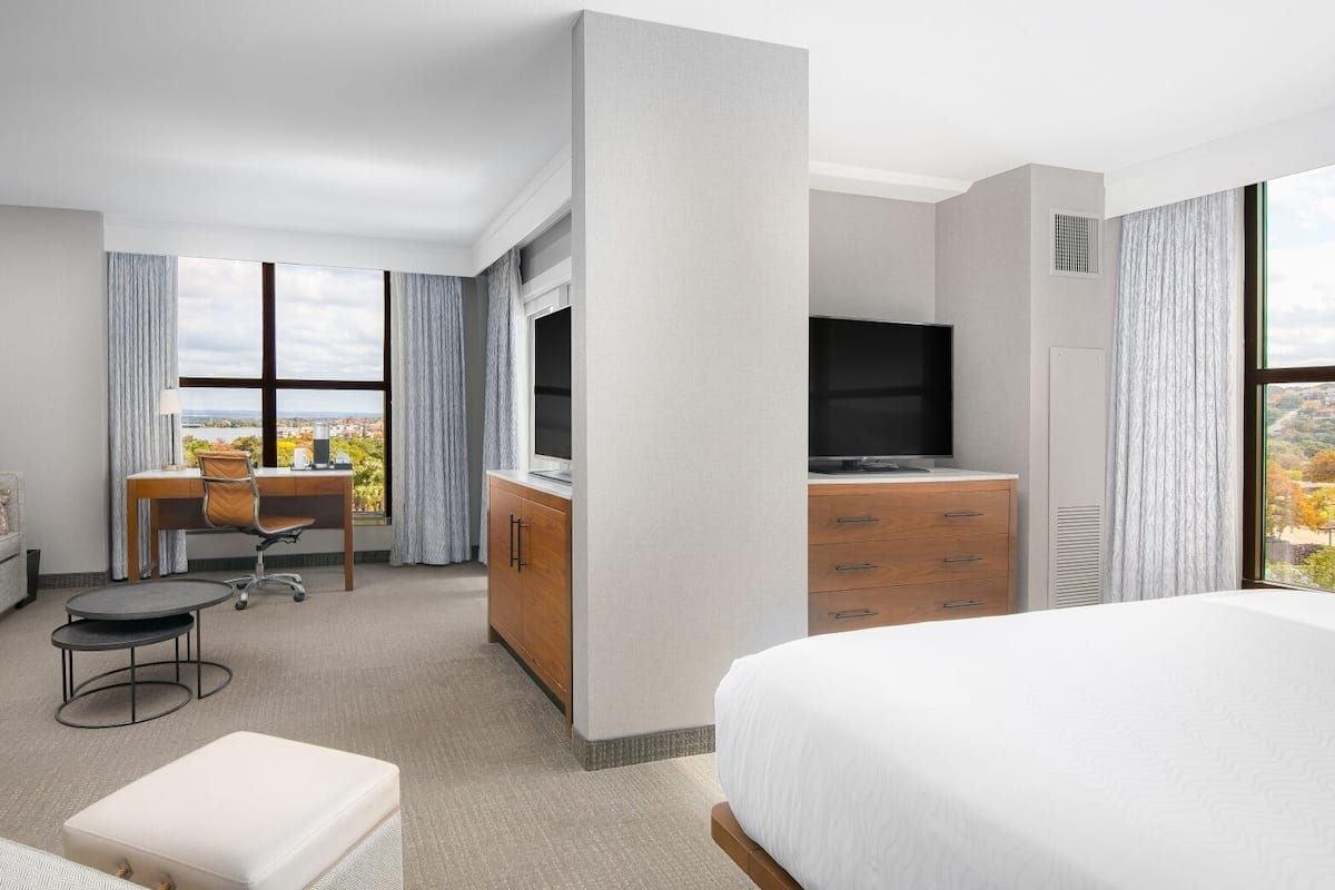 A hotel room with a bed, a desk, a sofa, a chair, a TV, and a window with a city view.