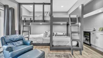 Habitación con literas, un sillón y reposapiés de cuero azul, una alfombra pequeña y una zona de cocina con armarios y un refrigerador.