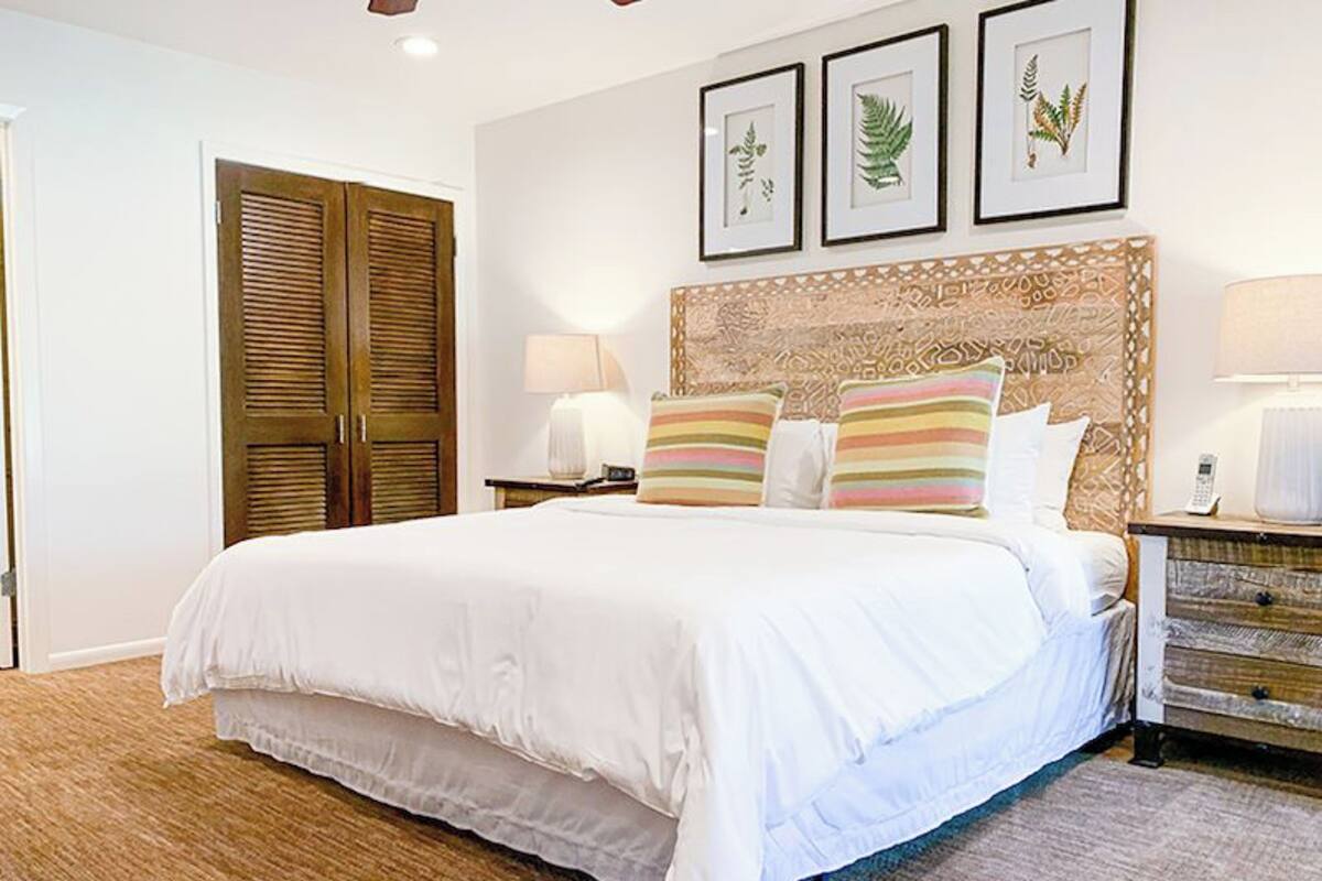A hotel room with a large bed, a wooden wardrobe, a nightstand with a lamp, and framed botanical prints on the wall.