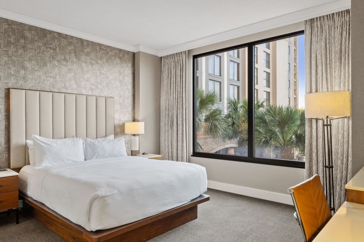 A hotel room with a large bed, two bedside tables with lamps, a view of palm trees and buildings through a window, and a wooden chair at a small table.