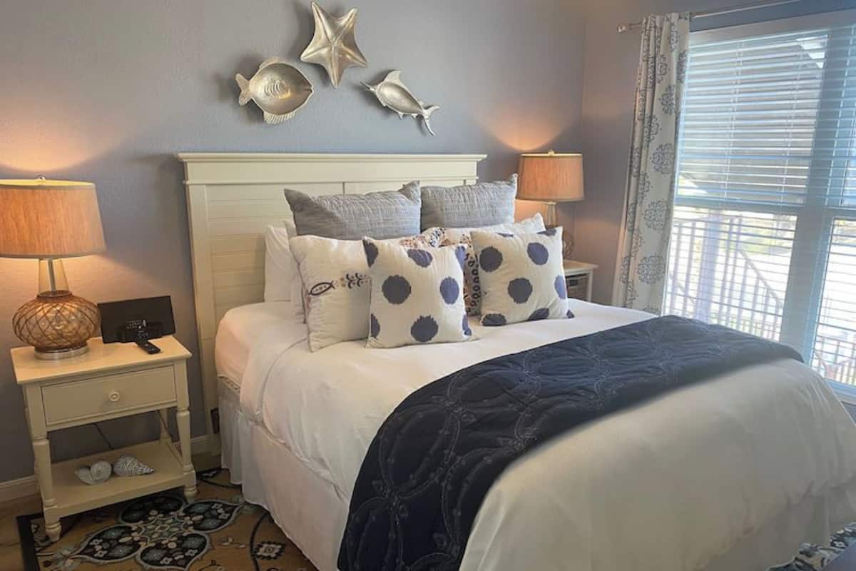 A neatly made bed with a dark blue bedspread, white pillows with blue polka dots, and a white headboard. There are decorative starfish on the wall, a lamp on the nightstand, and a patterned rug on the floor.