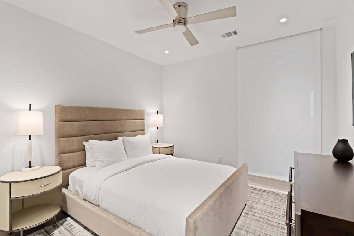 A modern hotel room with a large bed, two bedside tables with lamps, a ceiling fan, and a desk with a vase.